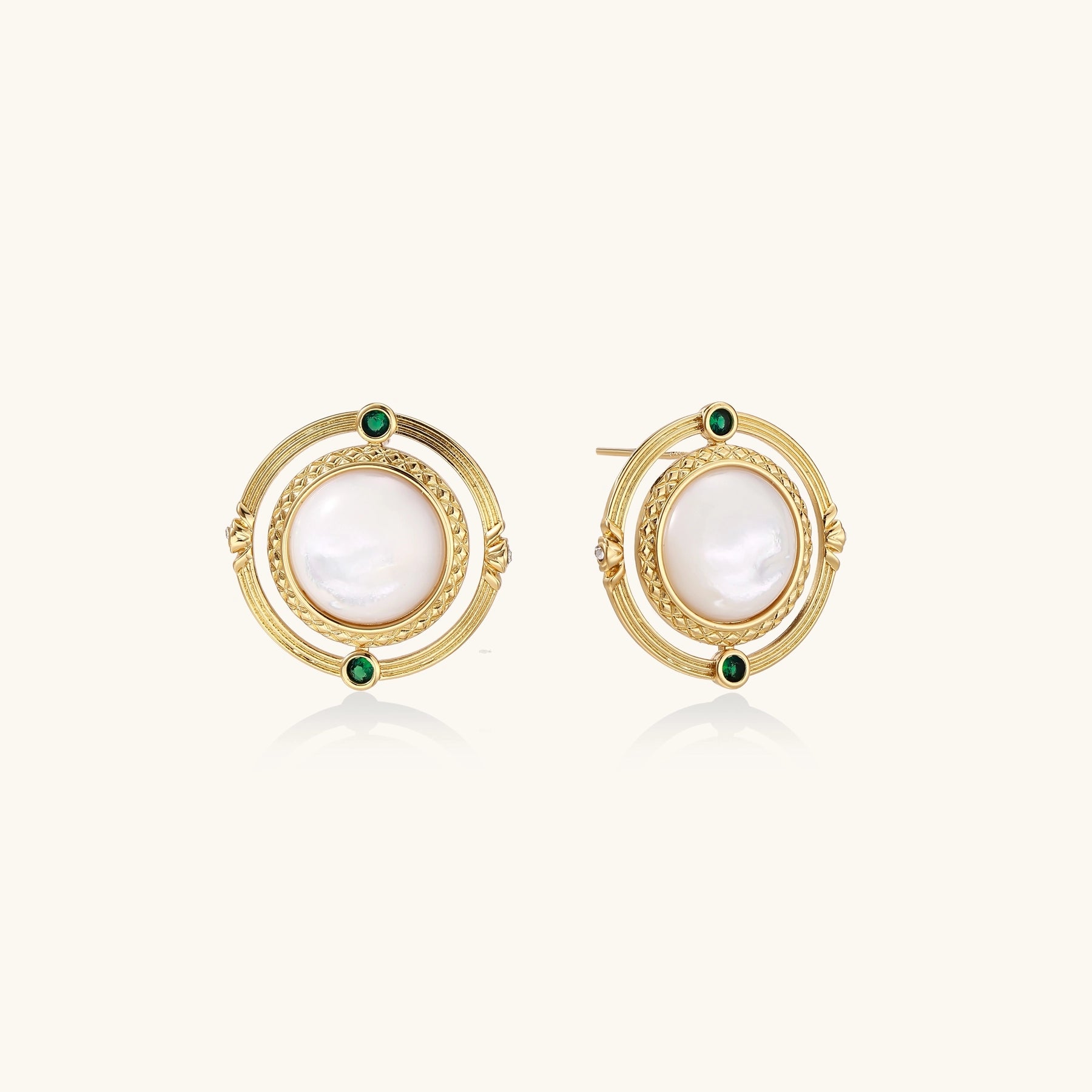 Close-up of gold-plated circular earrings with white mother-of-pearl centers and green zirconia accents on cream background.
