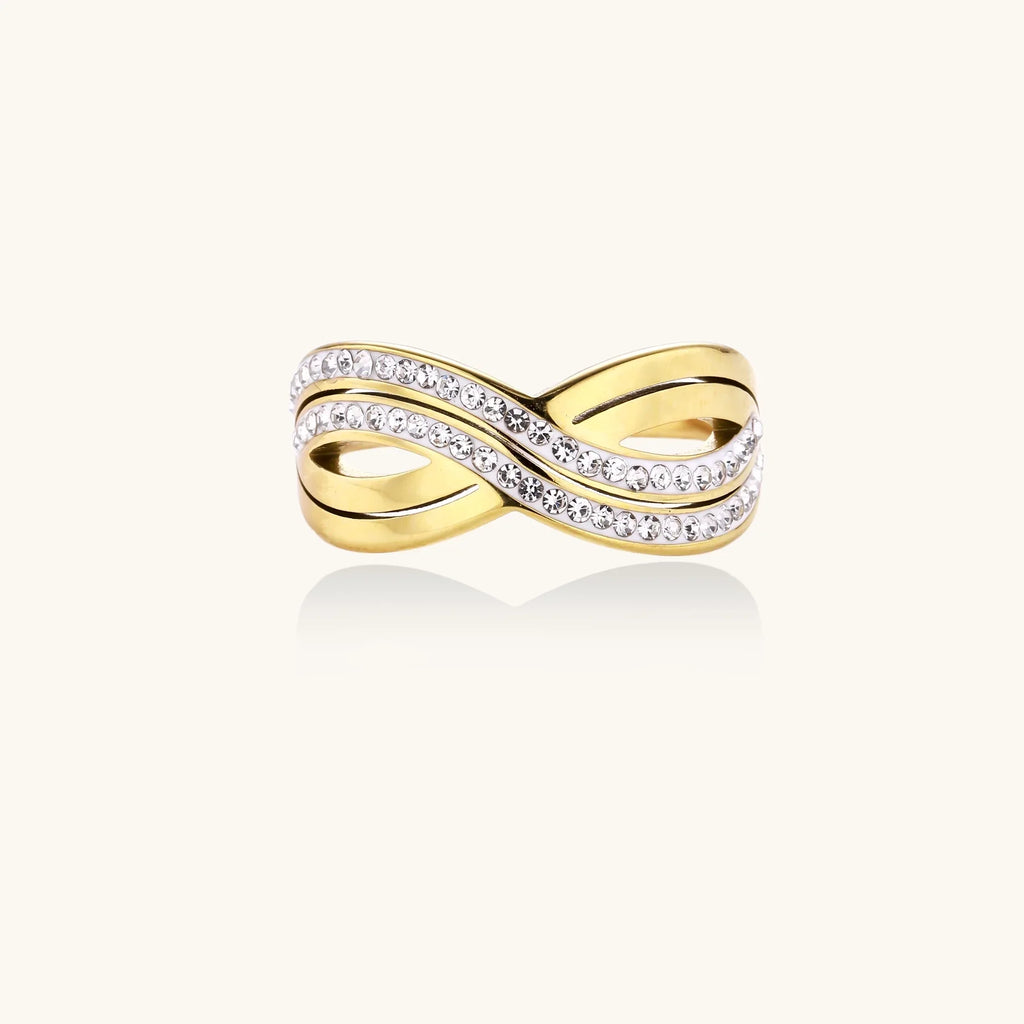 Gold-plated stainless steel ring with crossed infinity bands adorned with sparkling rhinestones against cream background.