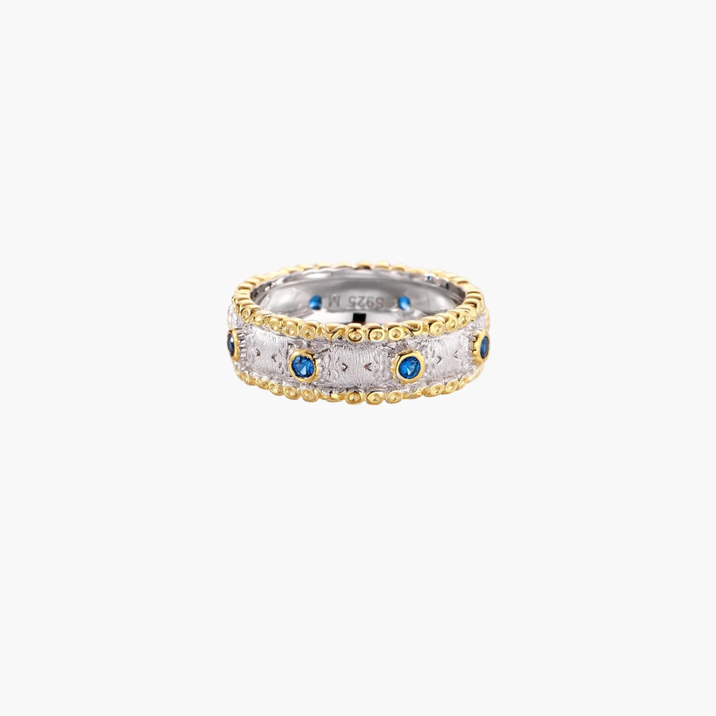 Two-tone ring with brushed silver center, gold beaded edges, and blue zirconia stones on white background.